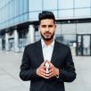 Portrait of a serious smiling modern Indian man, businessman, in front of the holding, behind him a modern building, he is looking at the camera.